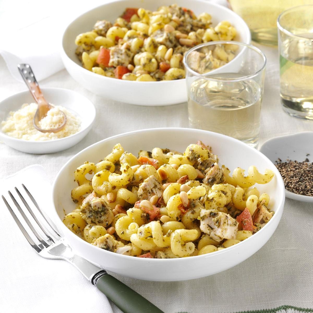 Pasta sits in a white bowl, mixed with chicken, pesto, and pine nuts; surrounded by a fork, Parmesan, pepper, and two glasses of white wine.