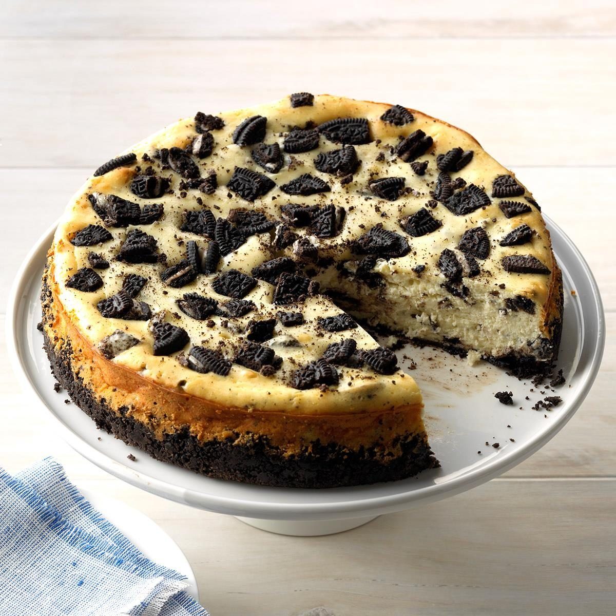 Cheesecake with Oreo pieces sits on a cake stand; a slice is removed, revealing a creamy interior in a bright kitchen setting.
