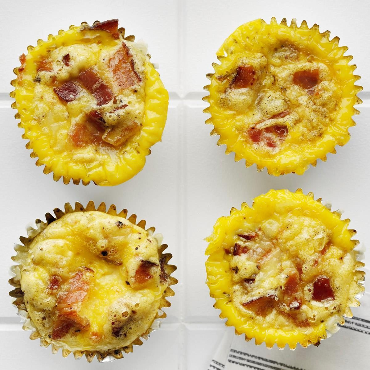 Egg muffins filled with bacon chunks rest in foil liners on a white tiled surface.