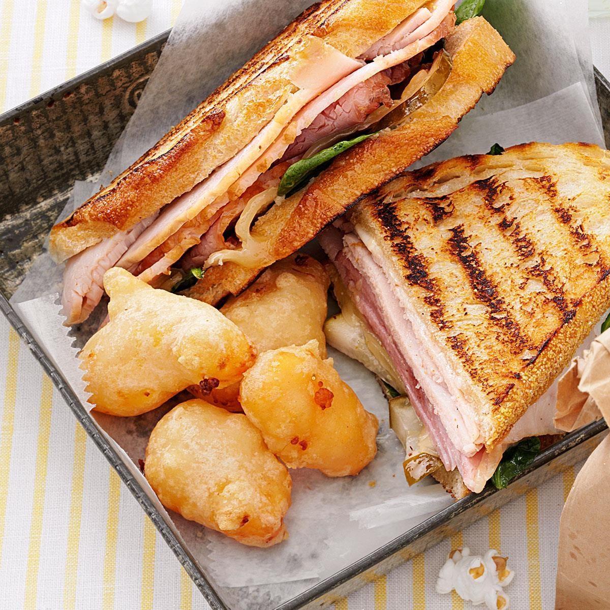 Grilled sandwich sits on parchment, surrounded by crispy fried cheese curds in a metal tray on a striped tablecloth.