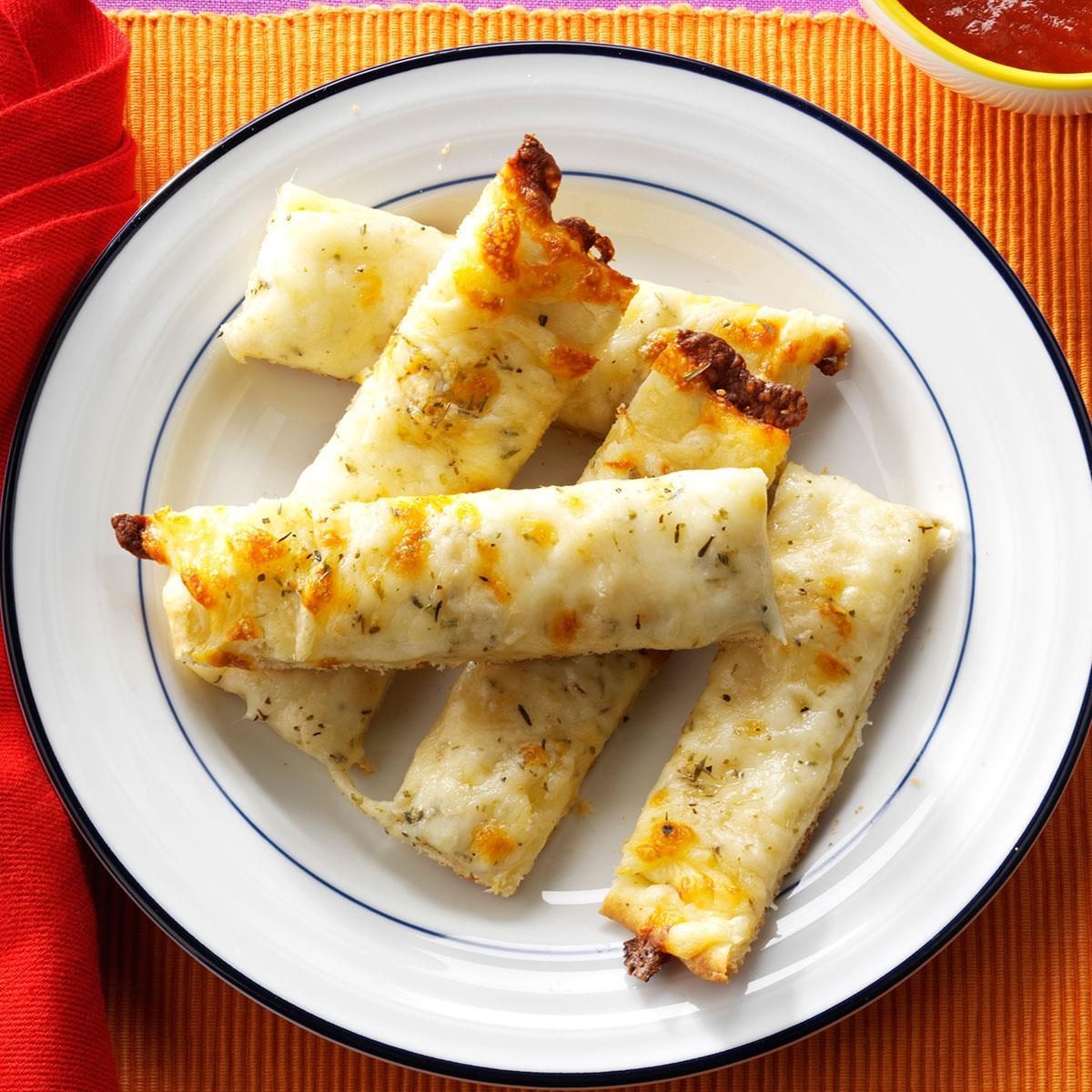 Cheese-covered breadsticks rest on a white plate with a blue rim, set on a colorful cloth beside a bowl of marinara sauce.