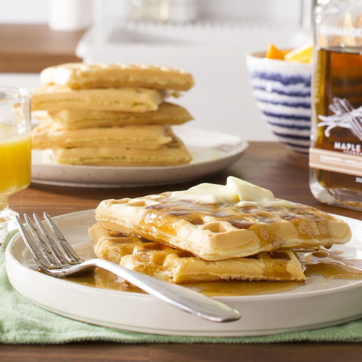 Waffles topped with syrup and butter rest on a plate with a fork; nearby are a syrup bottle, juice, and stacked waffles.