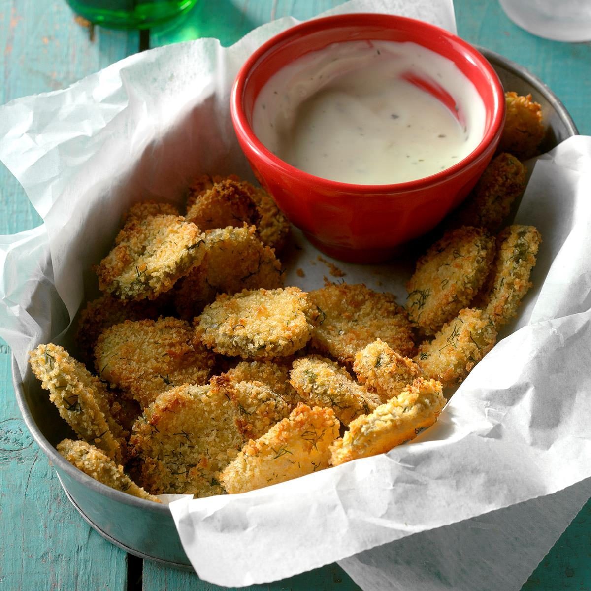 Breaded pickle slices rest in a bowl lined with parchment paper, accompanied by a red bowl of creamy dipping sauce on a turquoise wooden table.