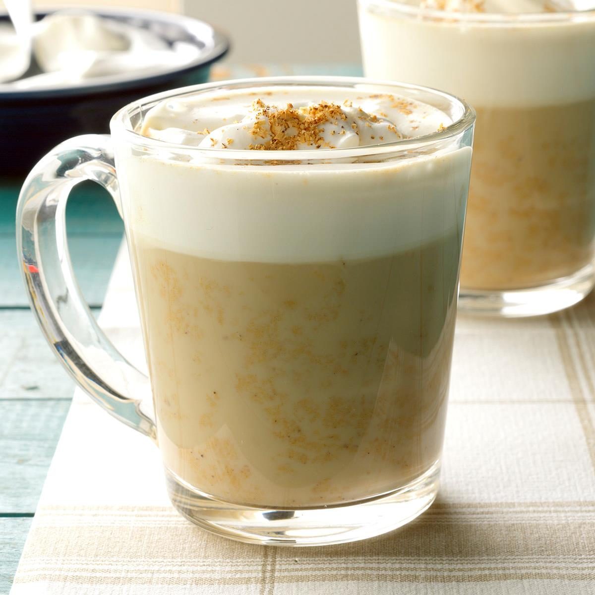 Clear mug containing frothy latte with whipped cream and cinnamon on top, placed on a striped placemat.