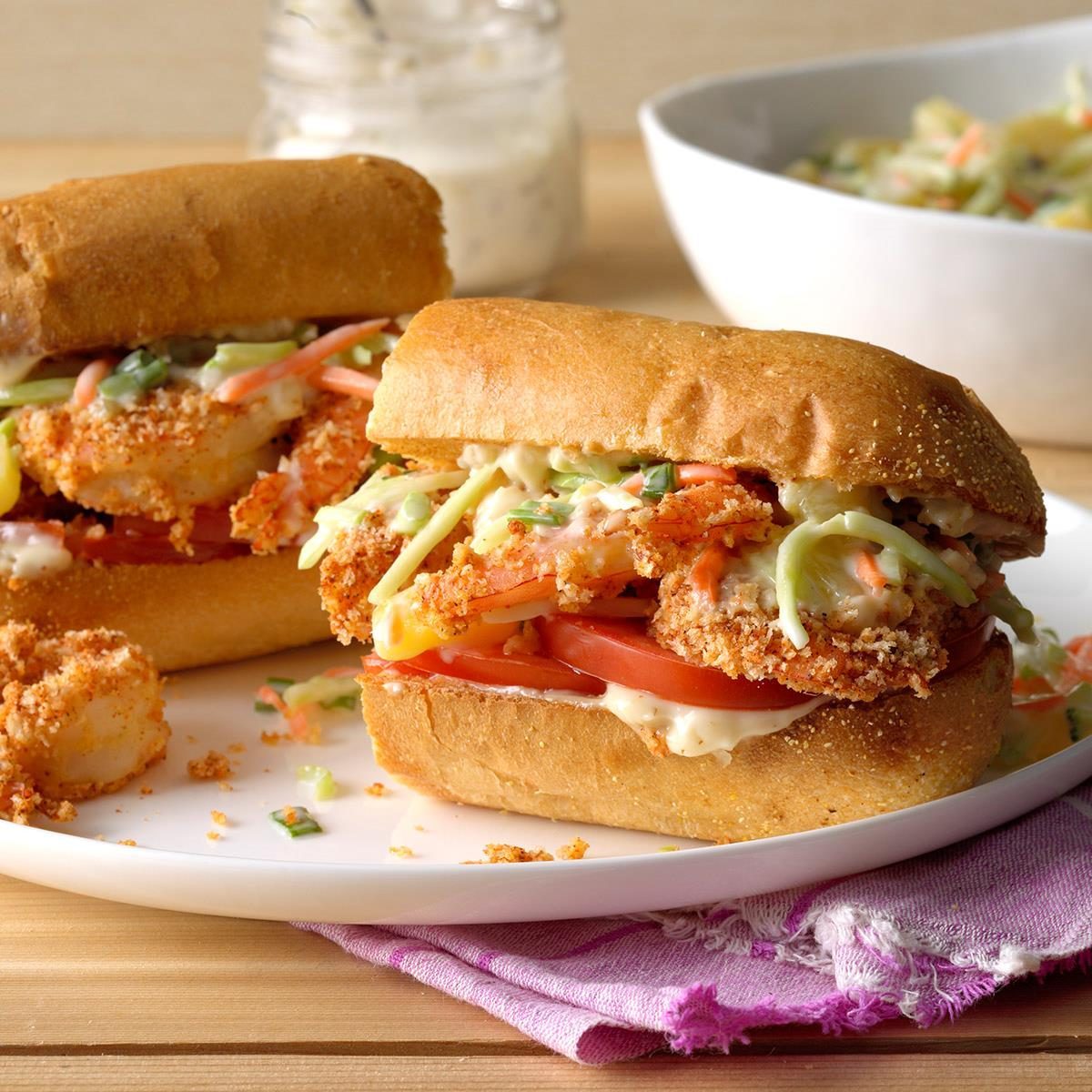 A sandwich filled with crispy shrimp and vegetables sits on a white plate. A jar of sauce and a bowl of coleslaw nearby.