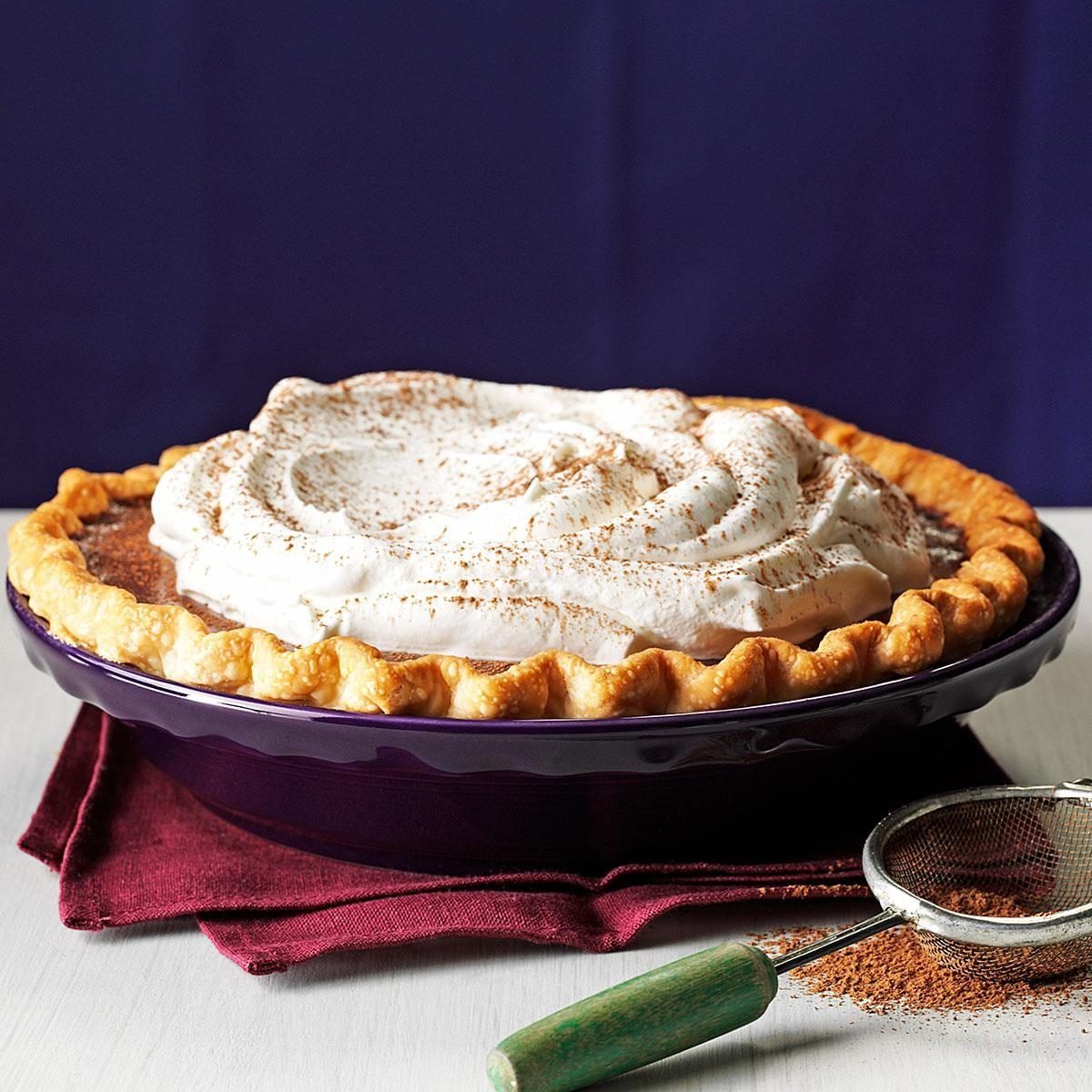 A pie topped with whipped cream rests in a purple dish on a red cloth, with a sifting tool nearby.