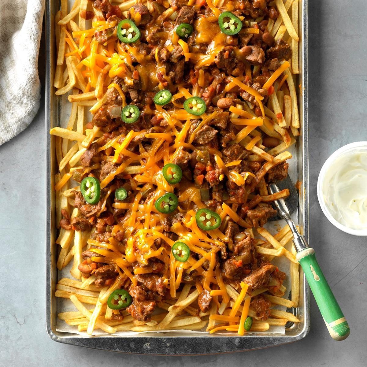 Fries topped with melted cheese, jalapeÃ±os, and meat on a baking tray, next to a bowl of sour cream on a gray surface.