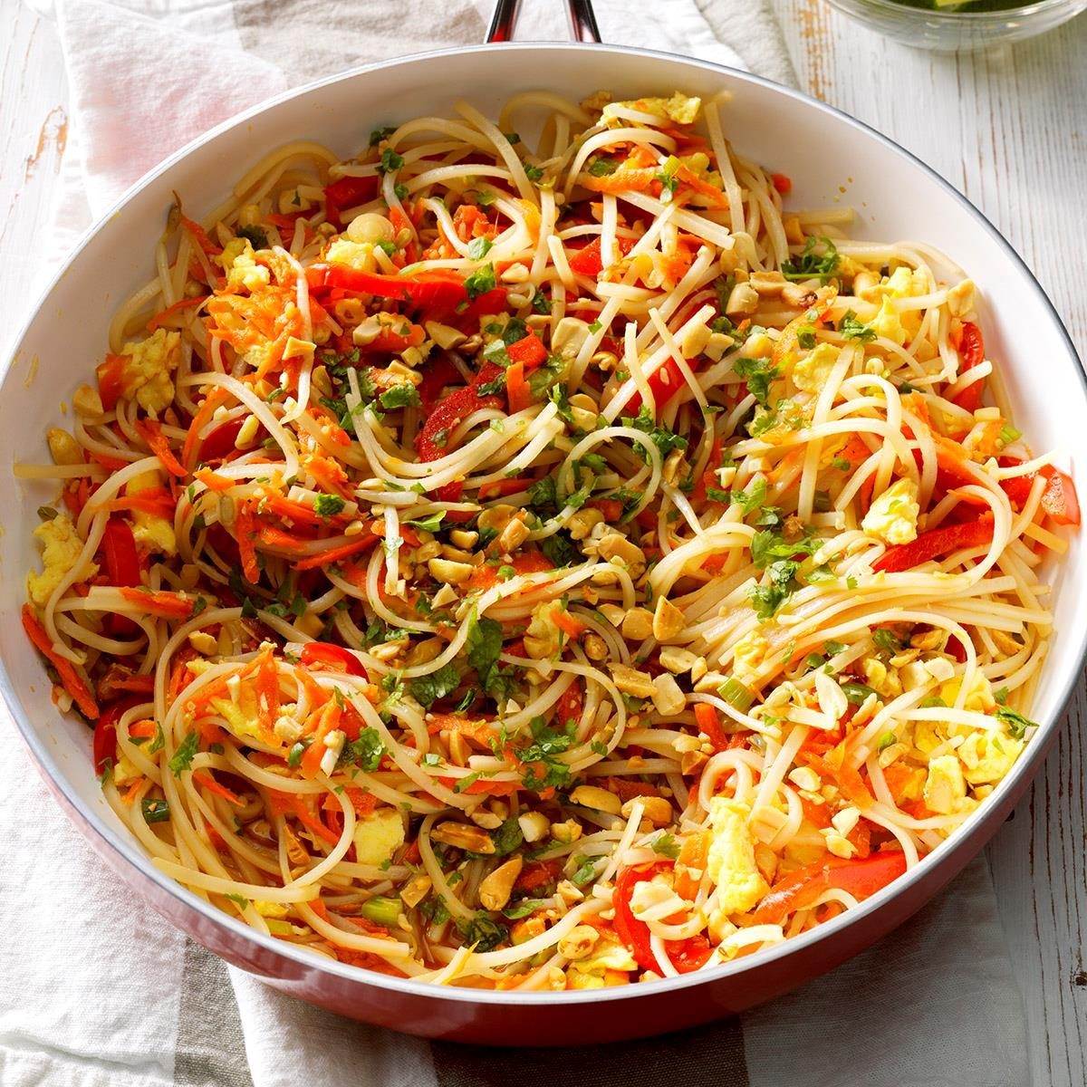 Noodles mixed with vegetables and nuts in a pan, resting on a wooden table with a cloth underneath.