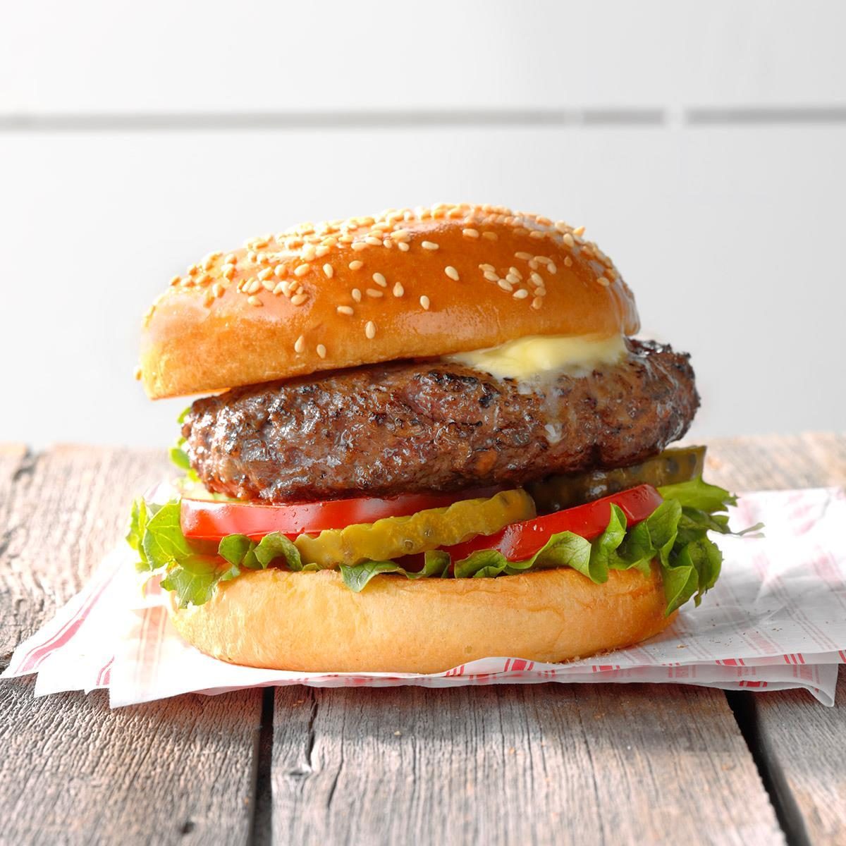 Burger rests on wooden surface, featuring a sesame bun, beef patty, cheese, lettuce, tomato, and pickles, against a light, blurred background.