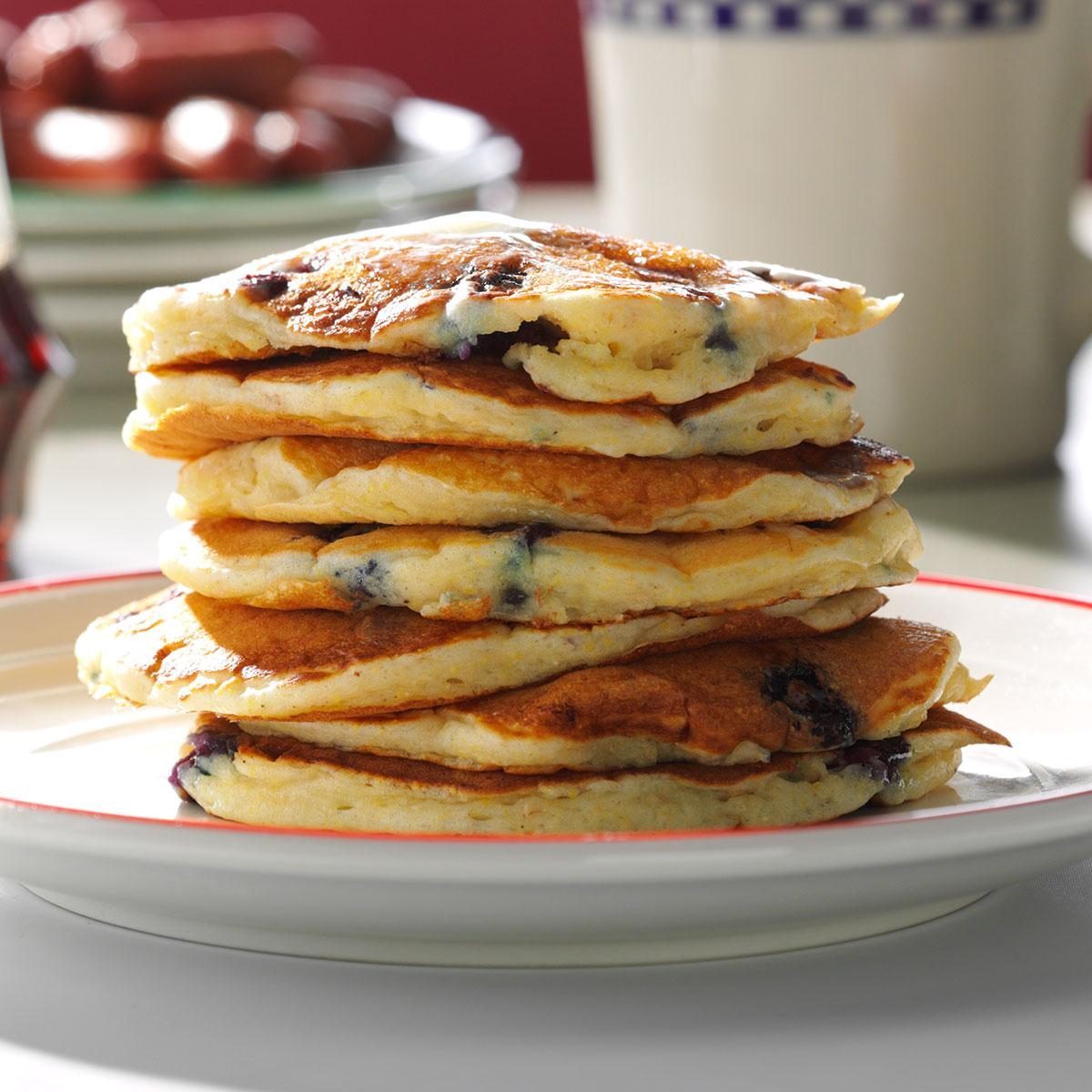 Stacked blueberry pancakes sit on a plate; a blurred background includes a mug and a distant dish.