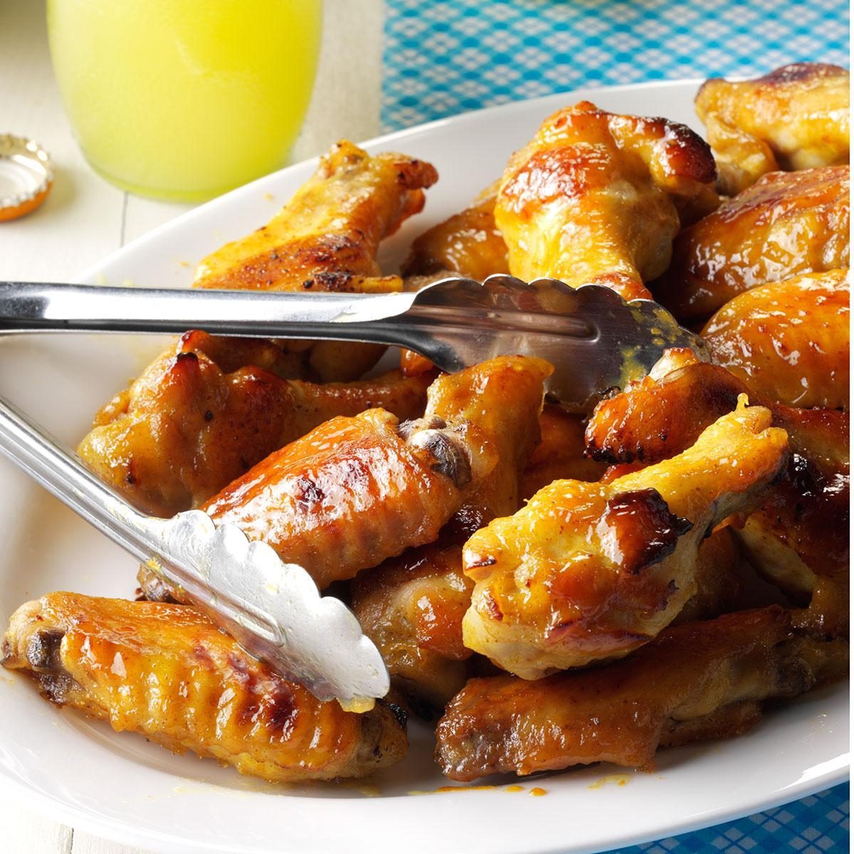 Chicken wings glistening with sauce; metal tongs grasping one piece. Background includes a glass of yellow liquid and blue checkered tablecloth.