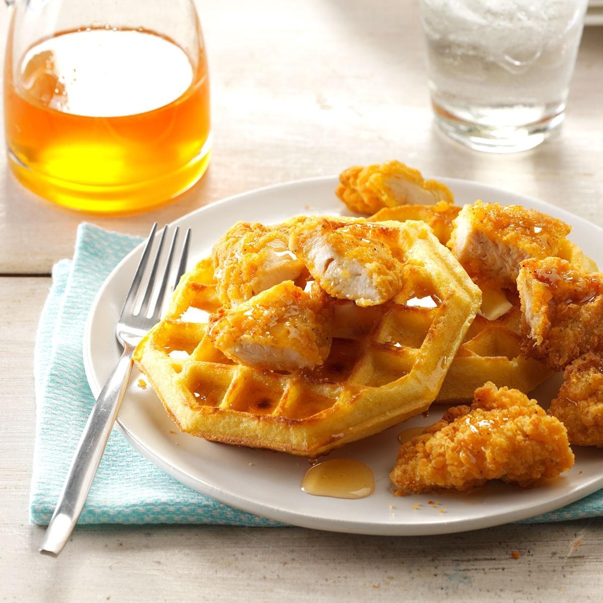 Fried chicken sits on waffles, drizzled with syrup, on a white plate with a fork; accompanied by a syrup jug and a glass of ice water.