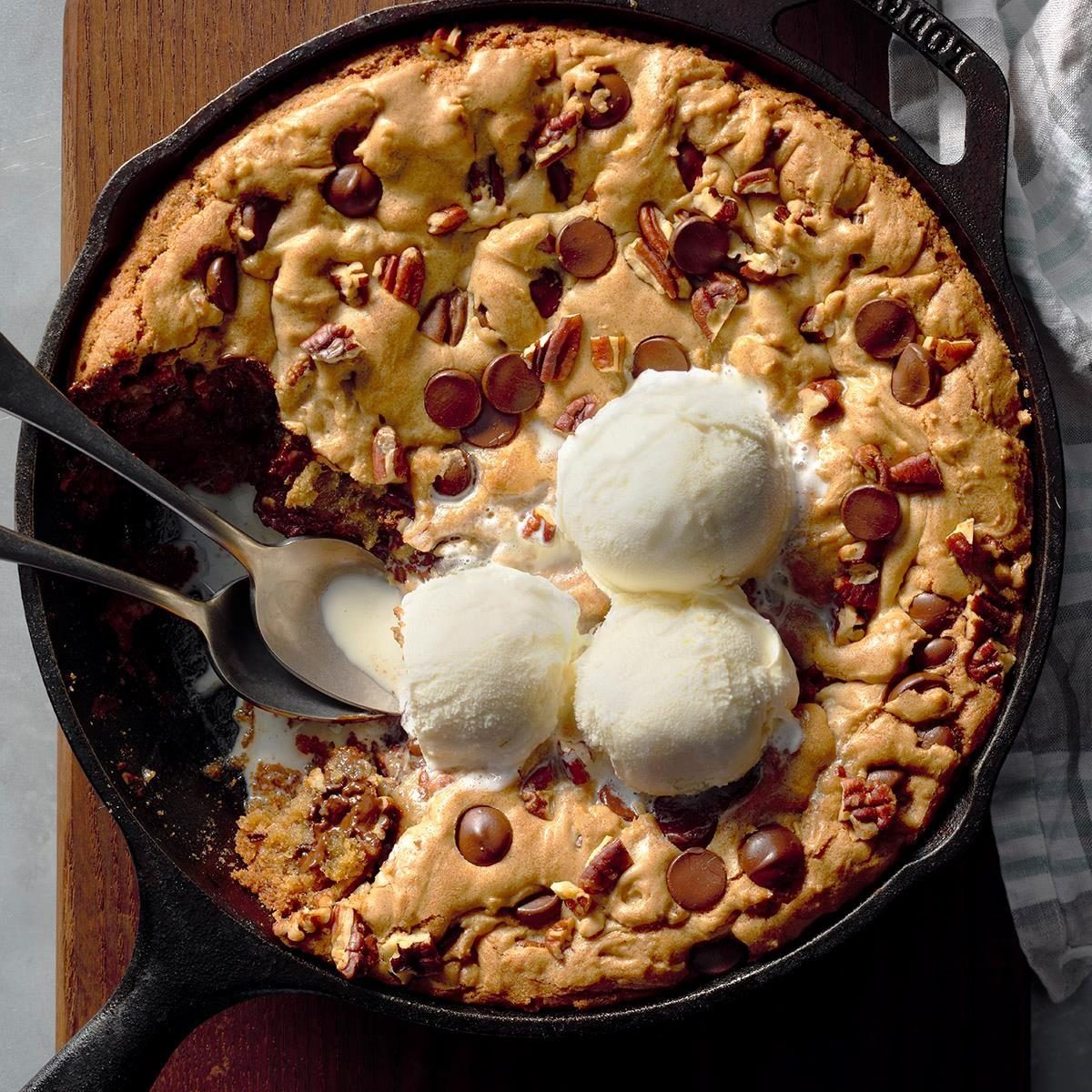 A skillet cookie topped with melting vanilla ice cream sits on a wooden board, surrounded by chocolate chips and pecans, with two spoons inserted.