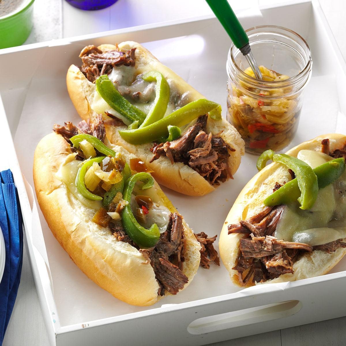 Sandwiches hold shredded beef and green peppers, served on a tray beside a jar of pickled vegetables on a white surface.