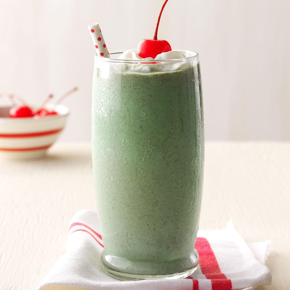 A green smoothie sits in a glass with whipped cream, a cherry, and a straw, placed on a cloth with a bowl nearby.