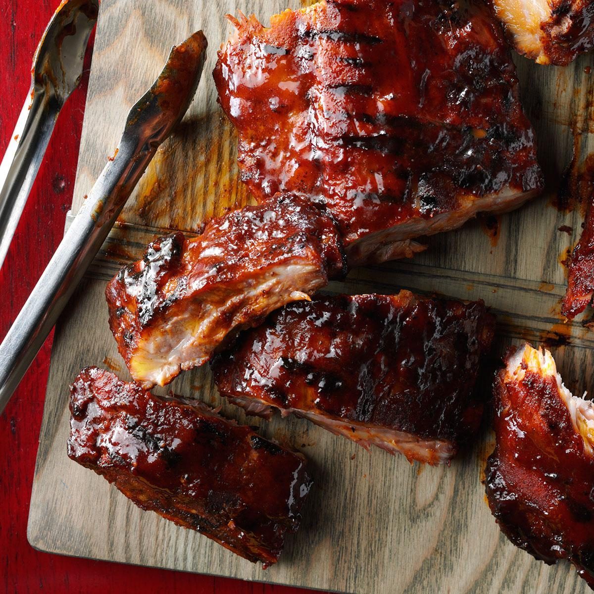 Barbecued ribs rest on a wooden board with metal tongs, surrounded by a red table.