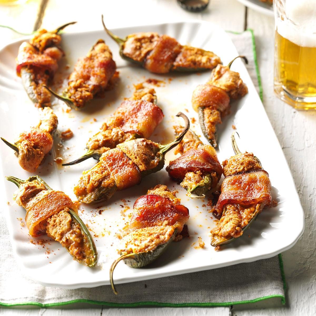 Bacon-wrapped stuffed jalapeÃ±os sit on a white plate, surrounded by a casual dining environment with a frothy glass of beer nearby.