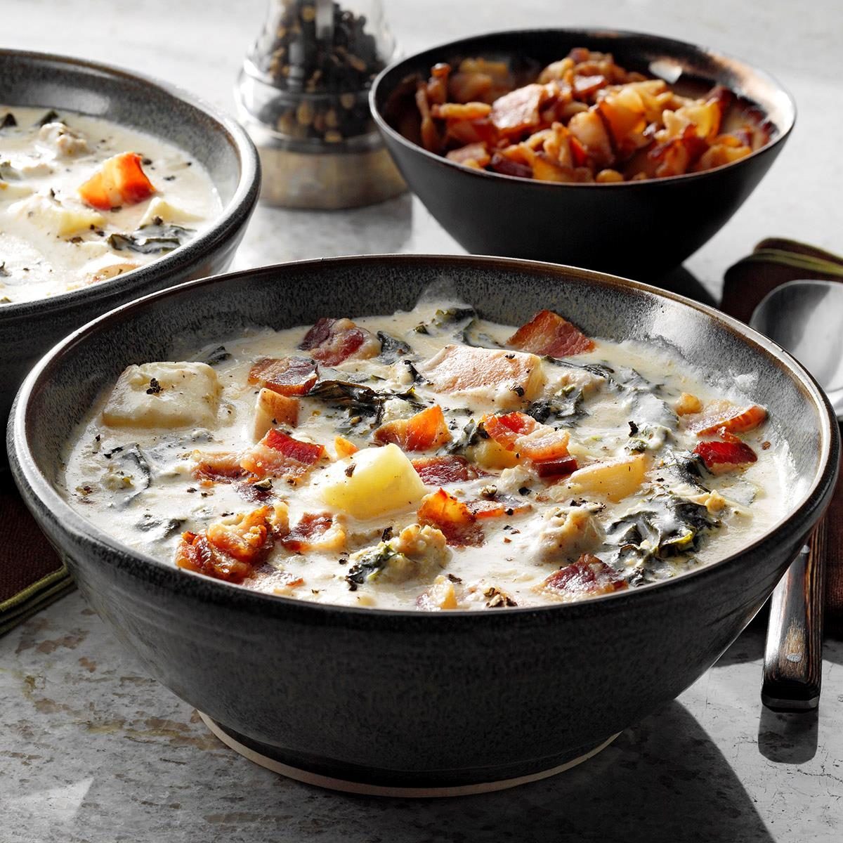 Bowl holds creamy soup with bacon and vegetables; other bowl contains extra bacon pieces; background shows a pepper grinder and cloth napkin.