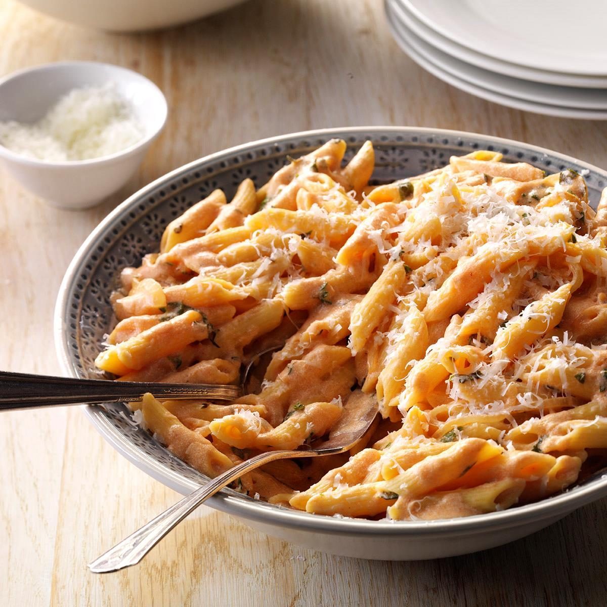 Pasta sits in a patterned bowl, covered in creamy sauce and shredded cheese, with a spoon and fork on a light wooden table.
