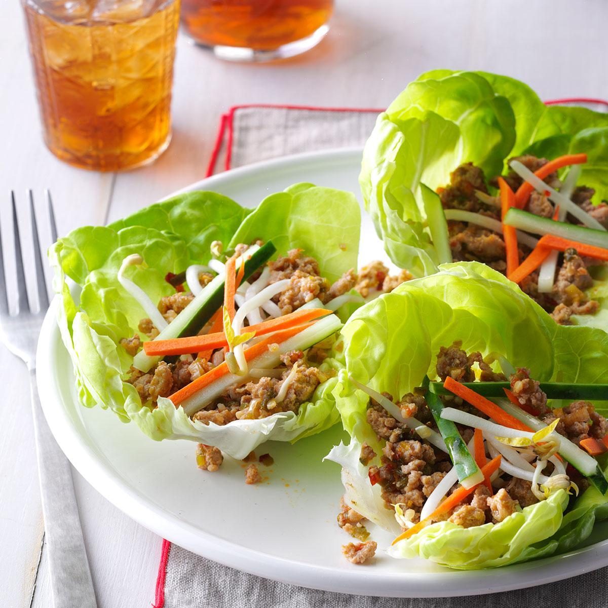 Lettuce cups hold ground meat, julienned carrots, cucumbers, and bean sprouts on a white plate with iced tea and a fork nearby.