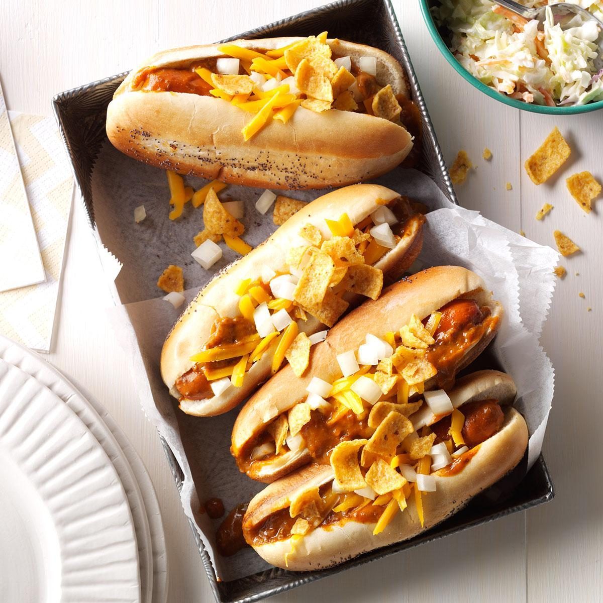 Hot dogs covered with chili, diced onions, cheddar cheese, and corn chips, resting in a tray with a bowl of coleslaw nearby.