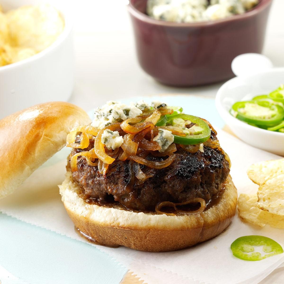 Burger topped with caramelized onions, blue cheese, and jalapeÃ±o slices, resting on a bun; nearby bowls contain chips and sliced jalapeÃ±os.