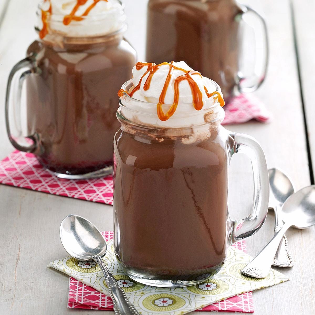 Glass mugs hold chocolate drinks topped with whipped cream and caramel drizzle, resting on colorful napkins with spoons nearby.