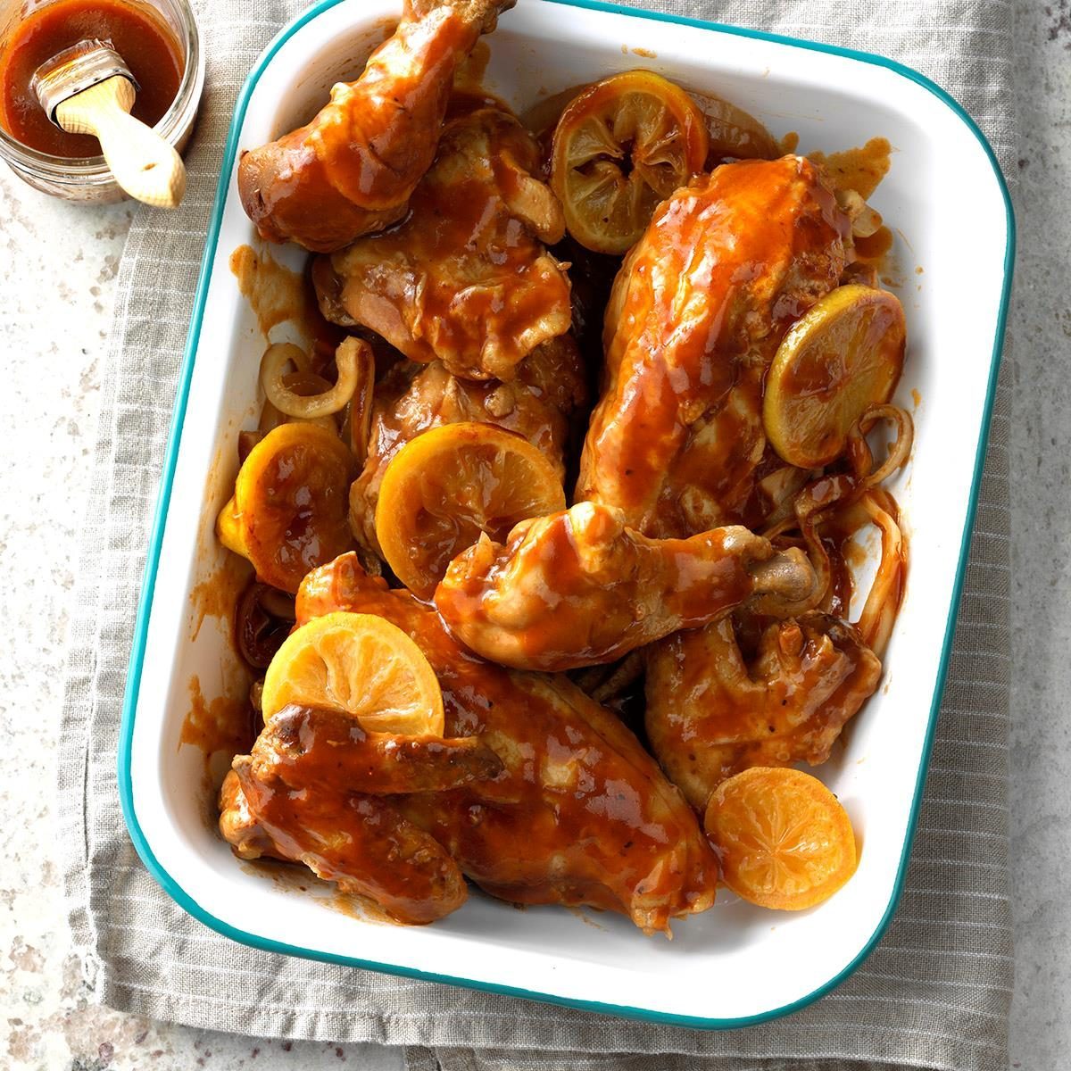 Chicken coated in barbecue sauce sits in a white dish with orange slices and onions, placed on a striped cloth near a jar of sauce.