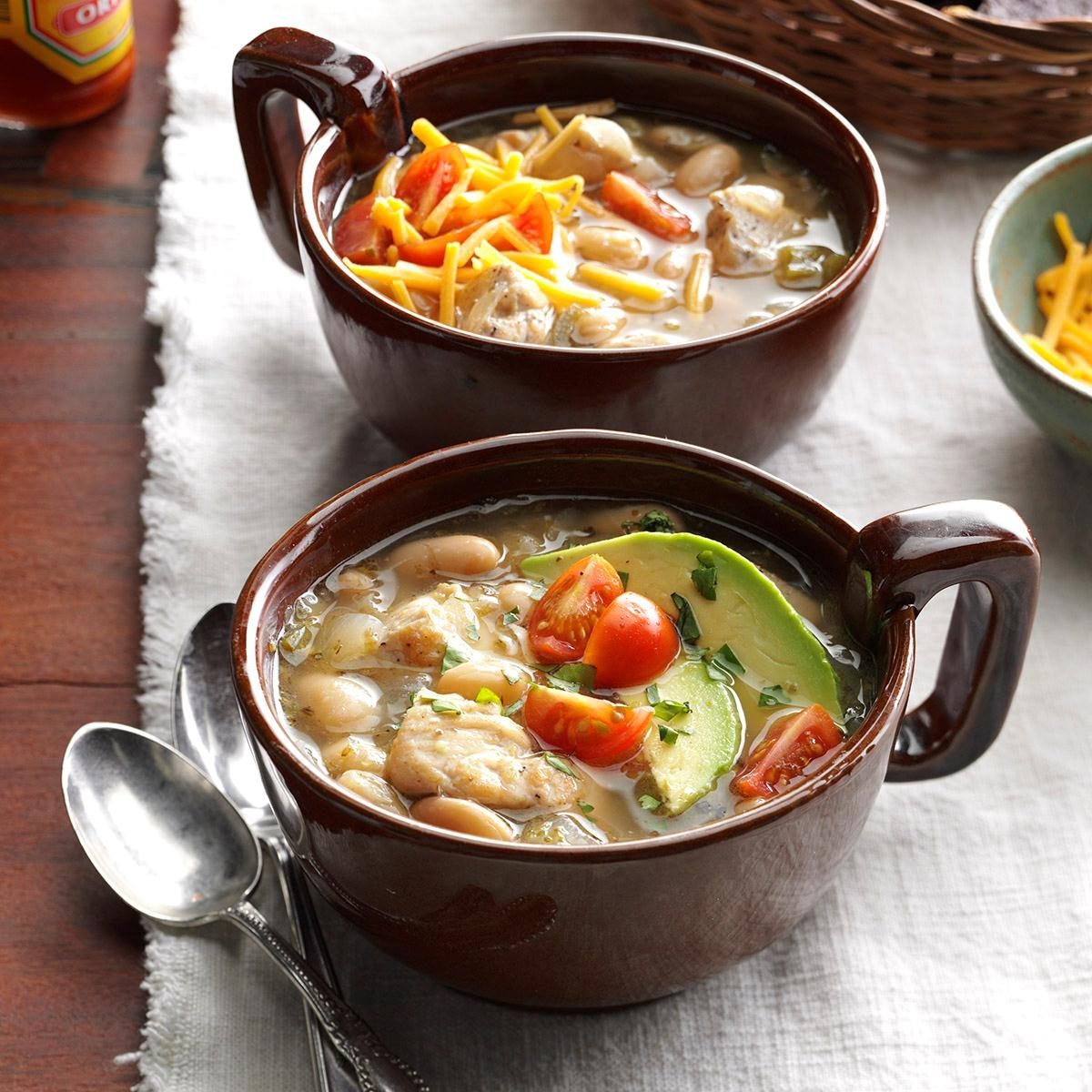 Two bowls of chili sit on a table, topped with cheese, avocado, and tomatoes, next to a basket and spoons.