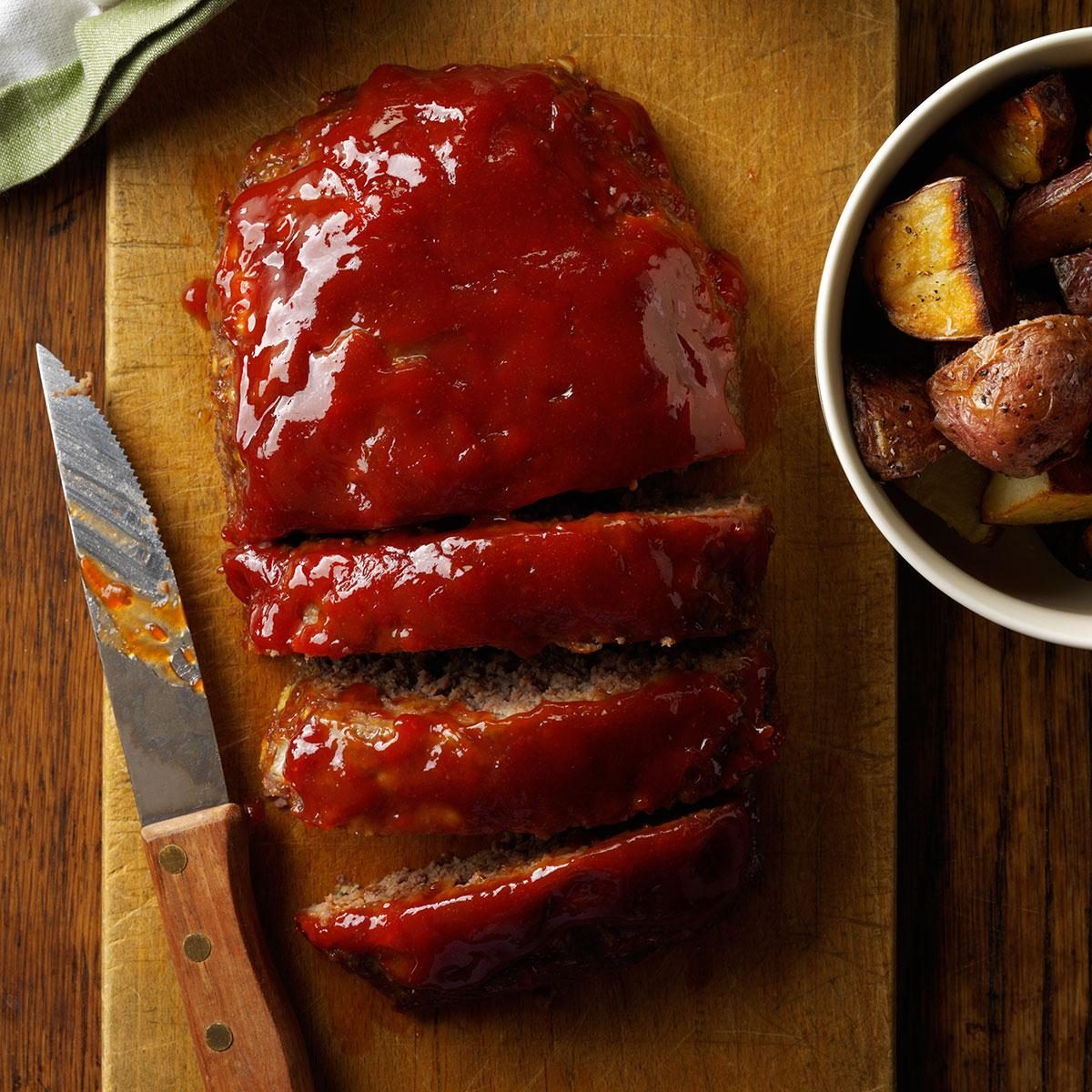 Sliced glazed meatloaf rests on wooden board beside a knife, with roasted potatoes in a bowl nearby.