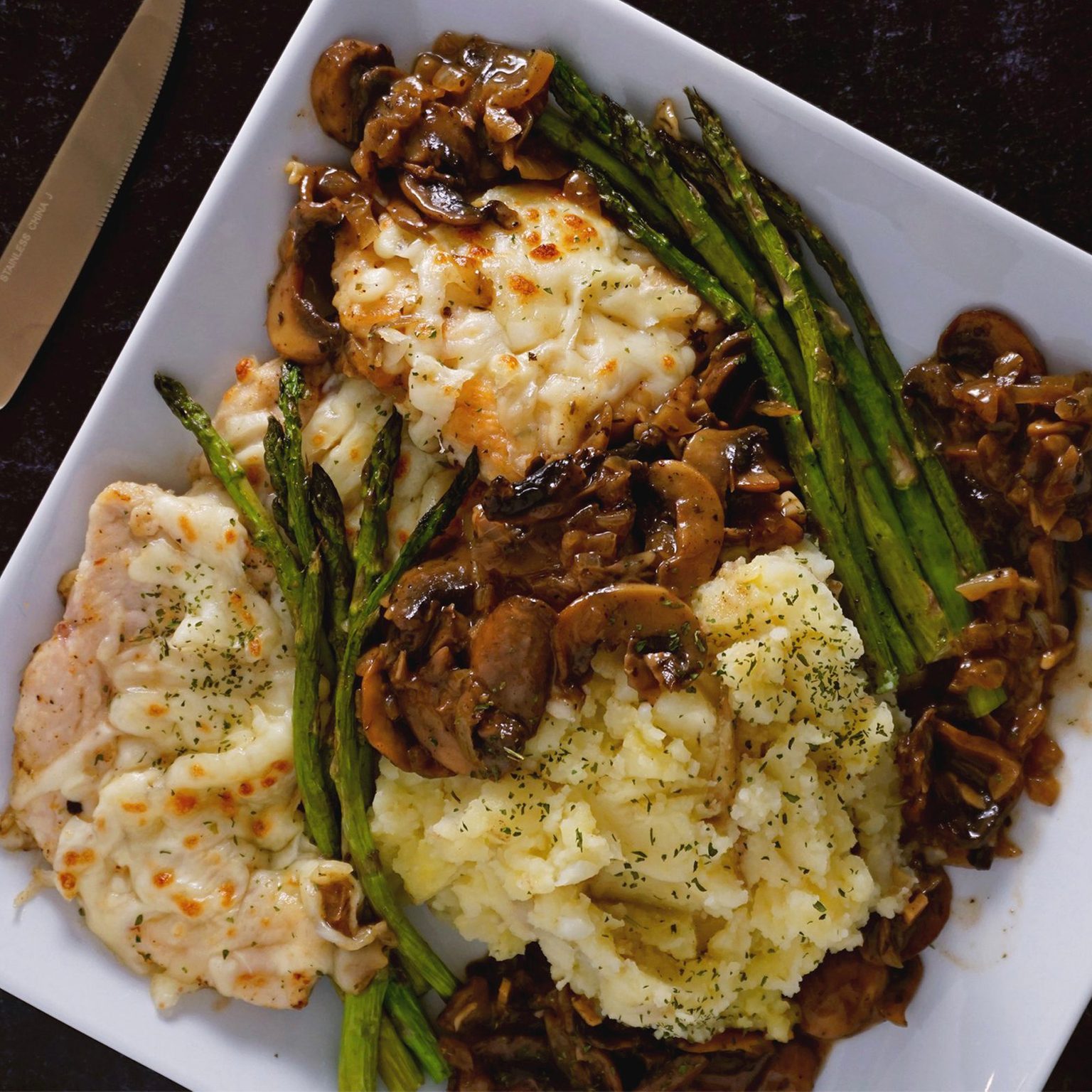 Chicken breasts covered in melted cheese rest on a plate with grilled asparagus and mashed potatoes, garnished with chopped herbs and sautÃ©ed mushrooms.