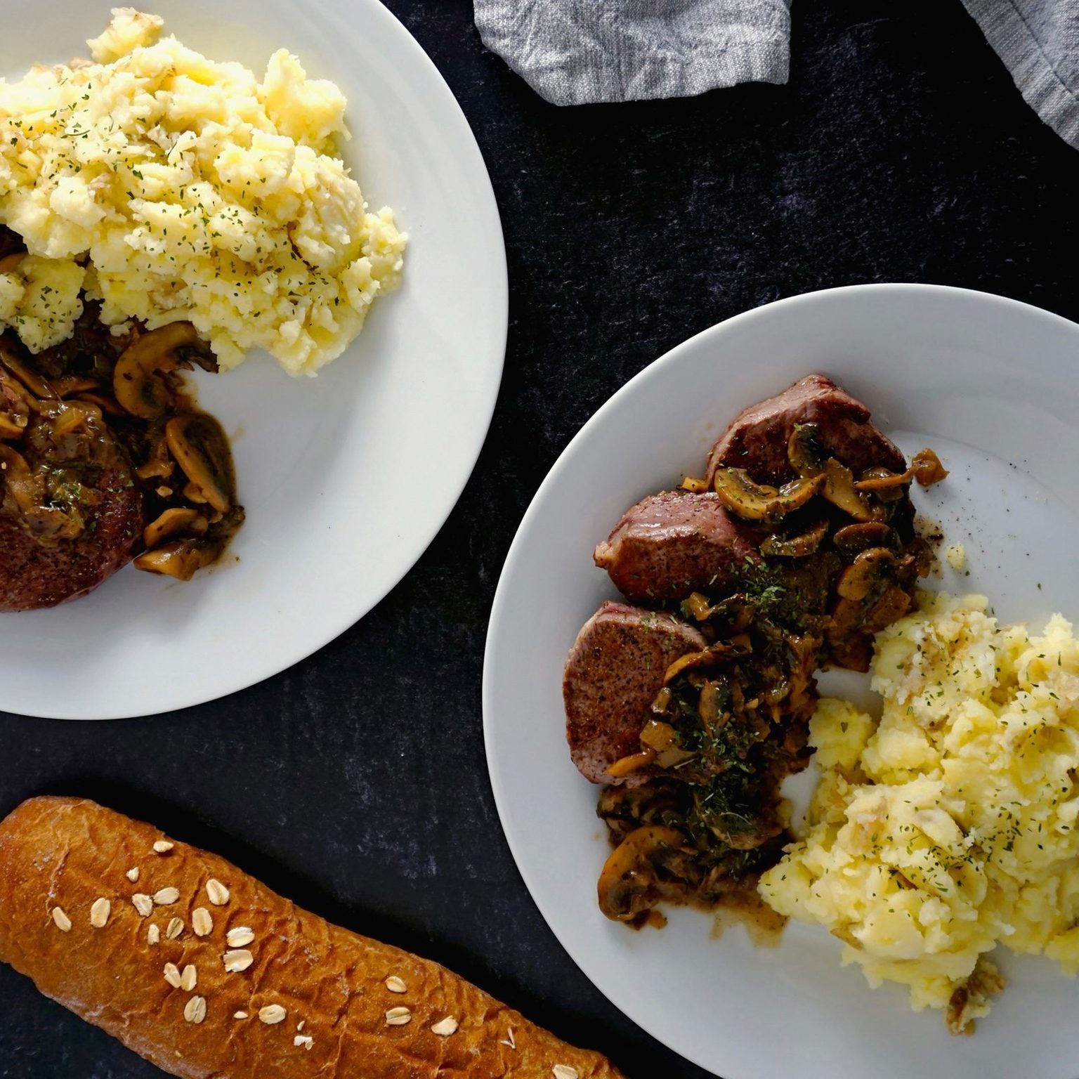 Plates hold steak with mushrooms and mashed potatoes, garnished with herbs; accompanied by a loaf of bread on a dark surface.