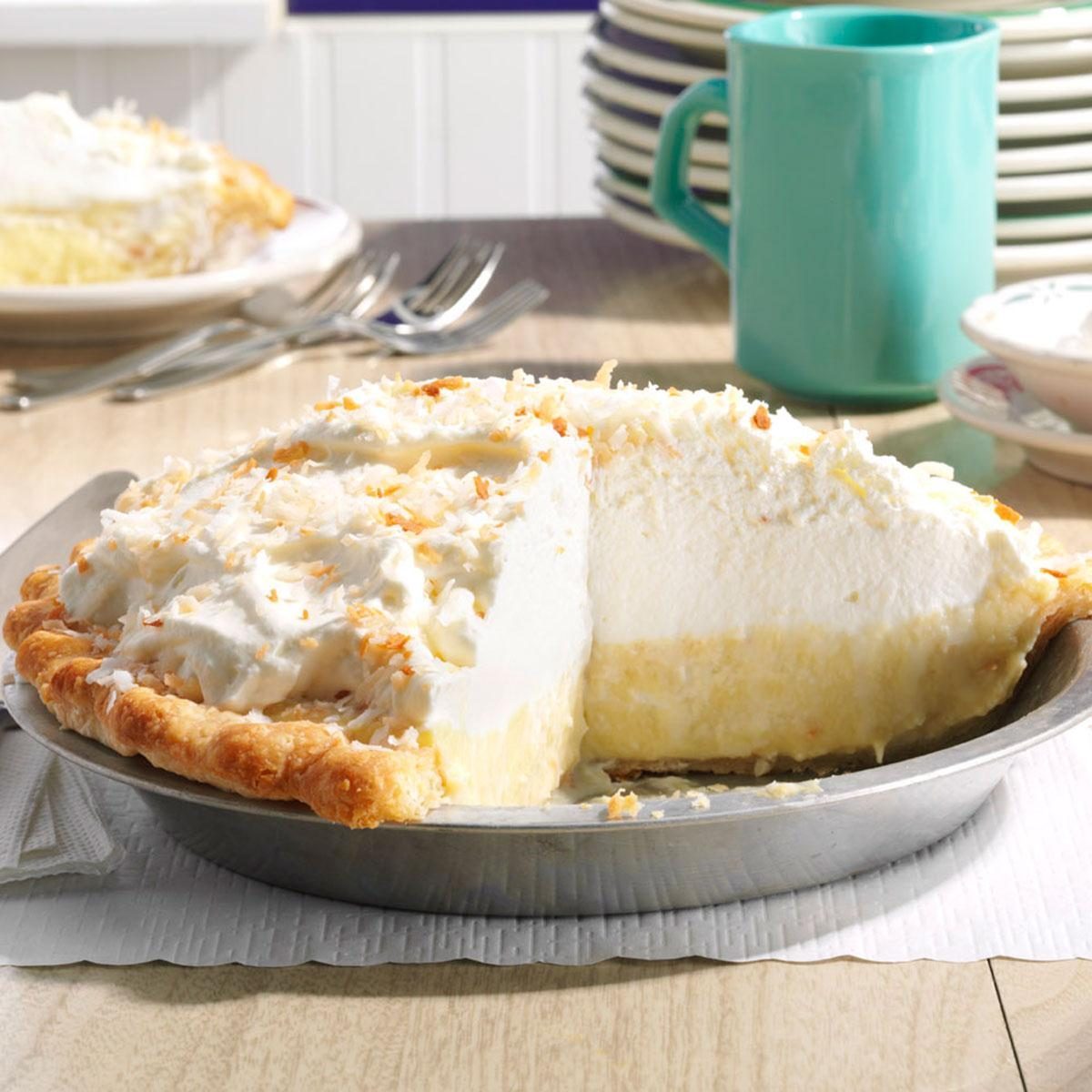 Cream pie sits with a slice removed, revealing layers, on a table with forks, a teal mug, and stacked plates nearby.