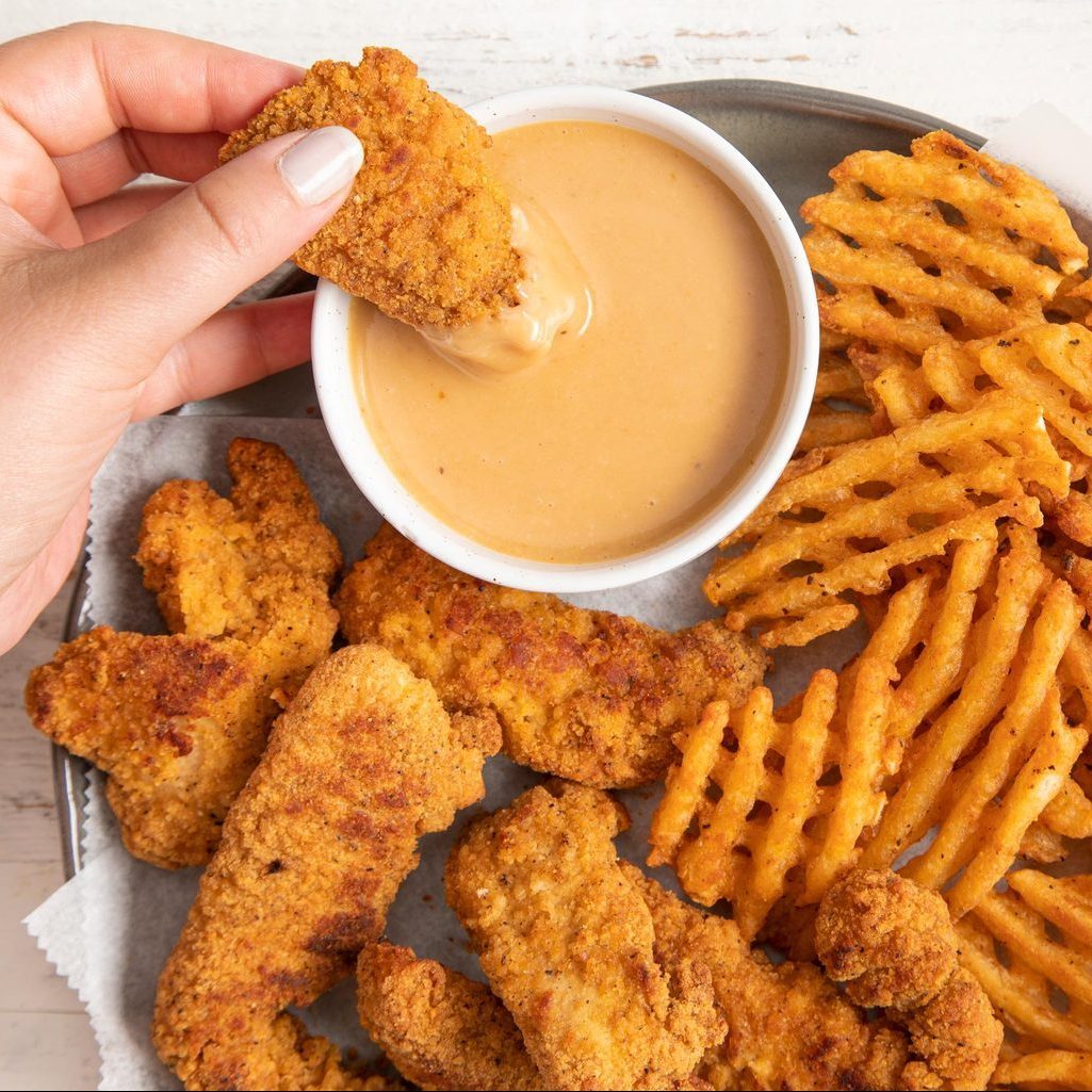 A hand dips crispy chicken tender into sauce, surrounded by waffle fries and more tenders on a metal plate with parchment paper.