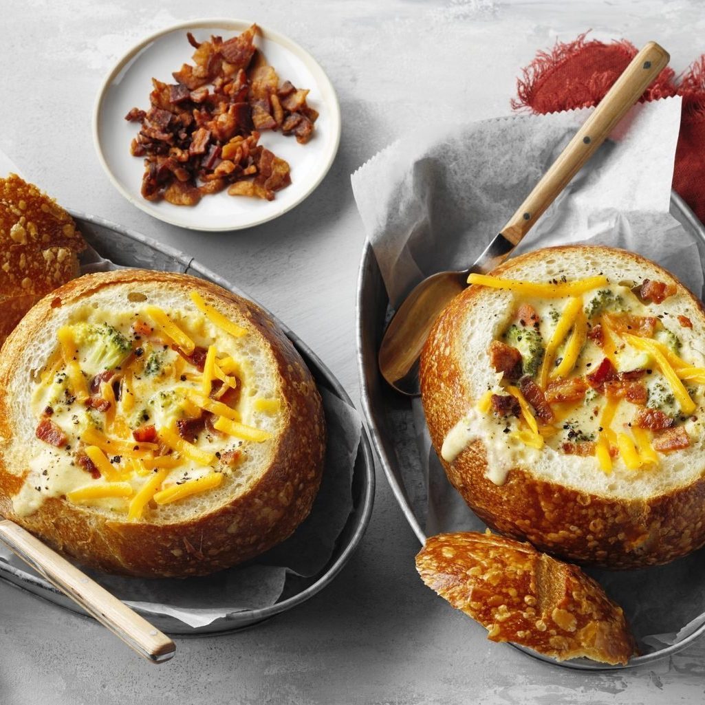 Bread bowls hold creamy soup topped with cheese and bacon; a small dish contains extra bacon. Served on metal trays with wooden spoons.