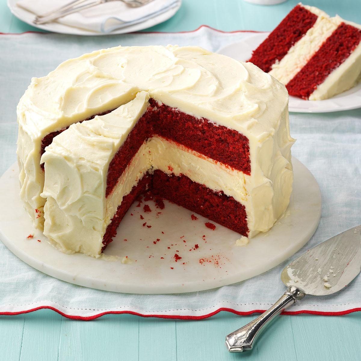 A red velvet cake with cream cheese frosting has a slice removed, sitting on a white plate atop a turquoise tablecloth.