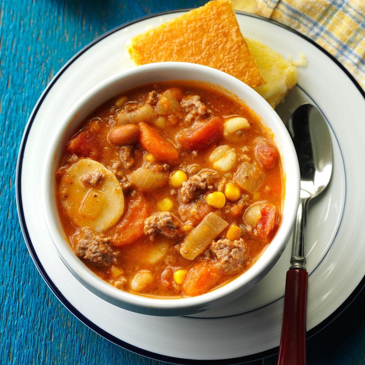 Bowl of chunky beef vegetable soup sits on a white plate, accompanied by a slice of cornbread and a spoon, on a blue table.