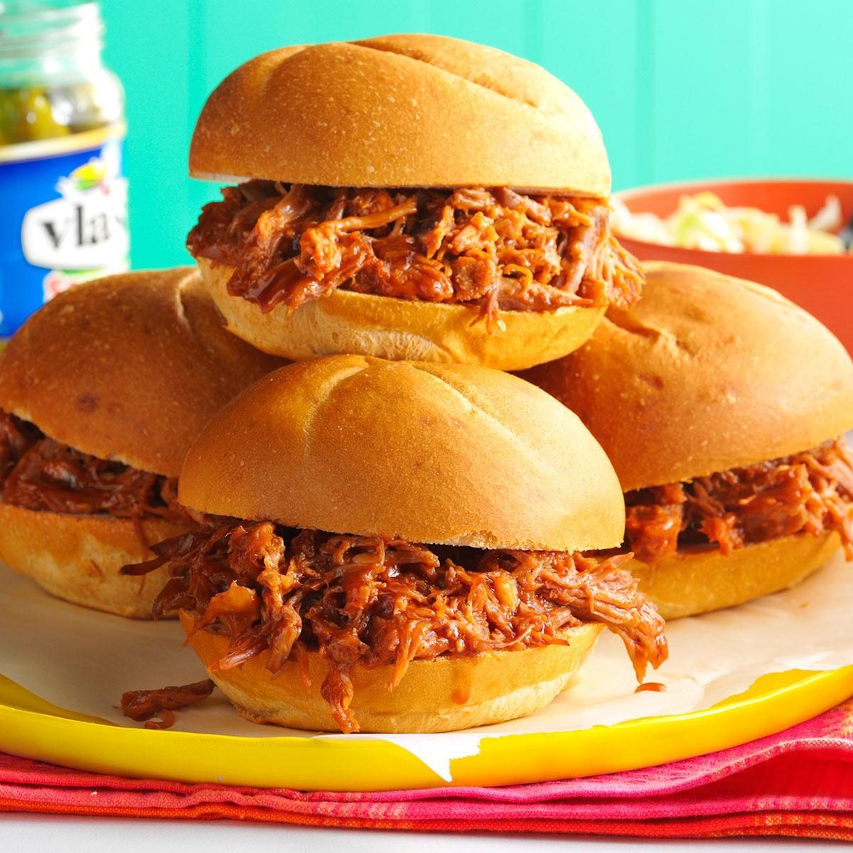 Sandwiches filled with shredded barbecue meat are stacked together on colorful napkins; a jar of pickles and a bowl are in the background.