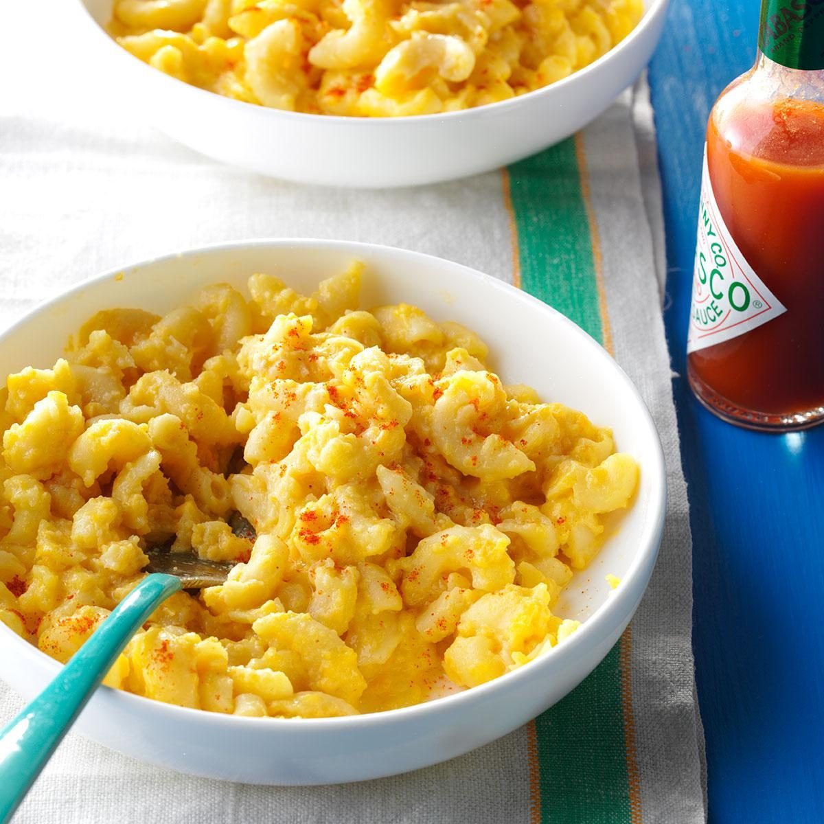 Macaroni and cheese sits in a white bowl with a fork; Tabasco sauce bottle on a colorful tablecloth.