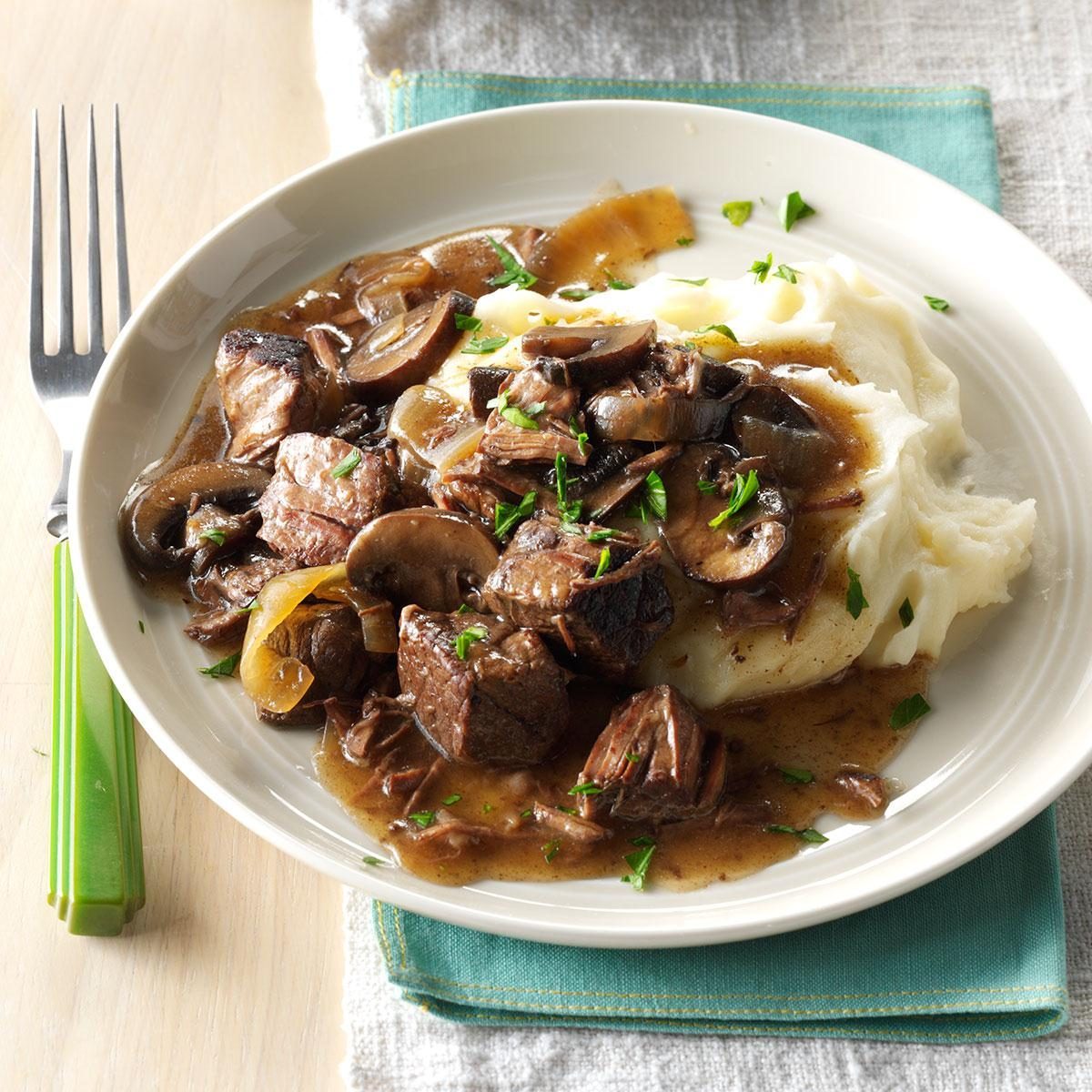 A plate holds beef and mushroom stew over mashed potatoes, garnished with herbs, beside a fork on a napkin-covered table.