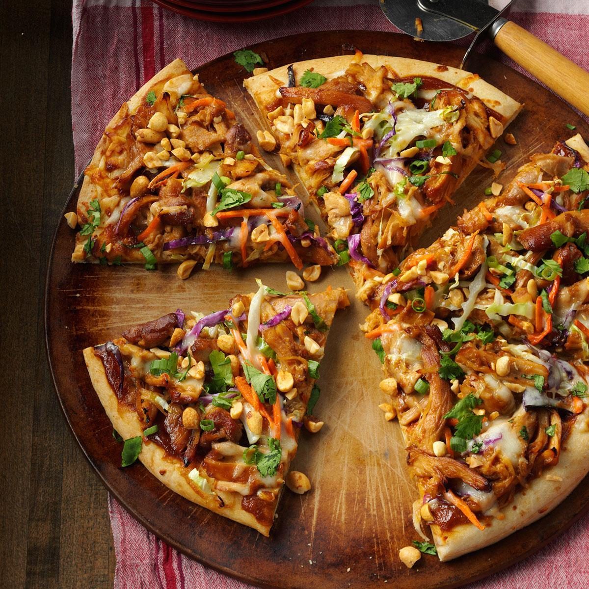 Pizza topped with chicken, peanuts, vegetables, and herbs on a wooden board, accompanied by a pizza cutter, set on a red-striped cloth.