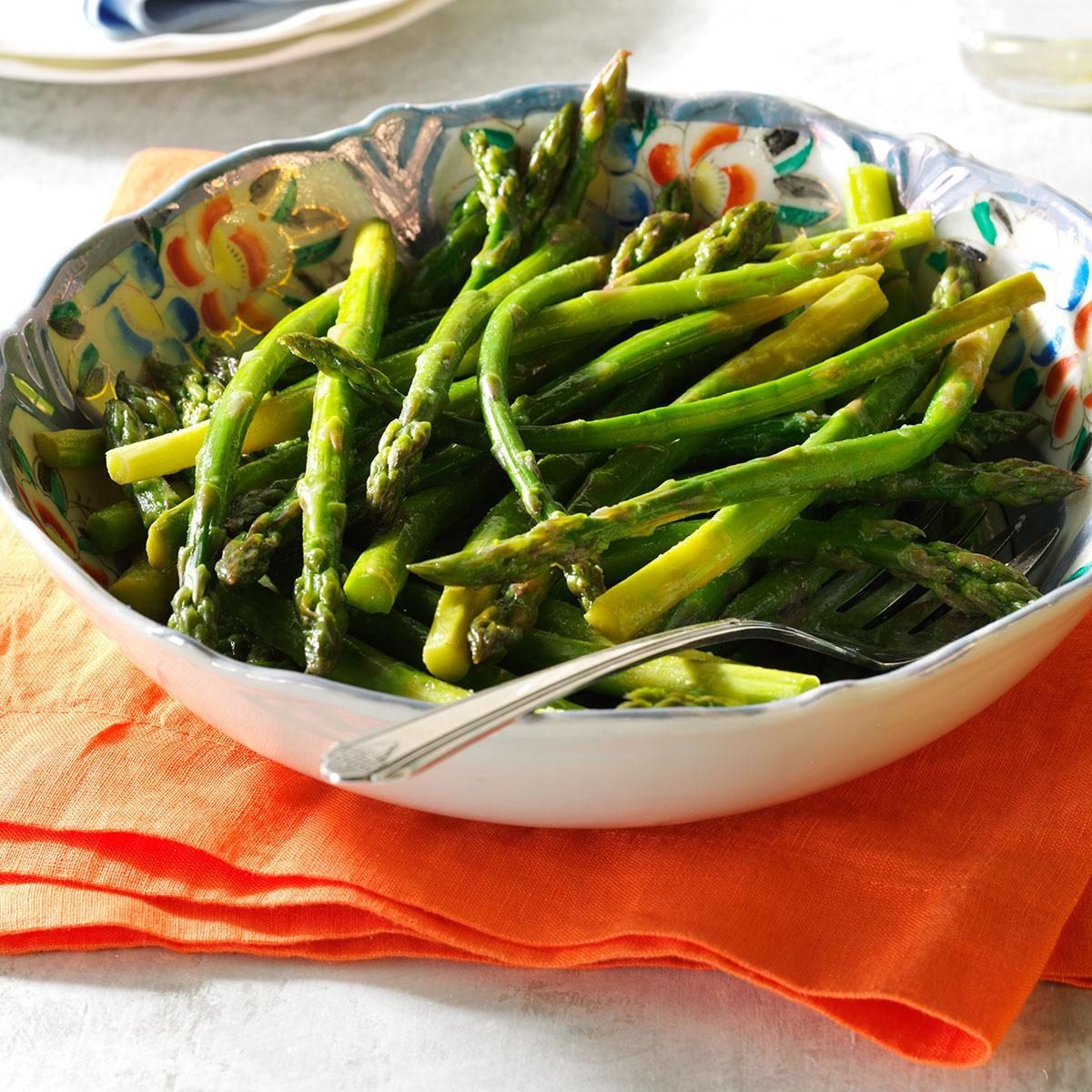 Asparagus sits in a decorative bowl on an orange napkin, with a fork resting beside it. Plates are visible in the background.