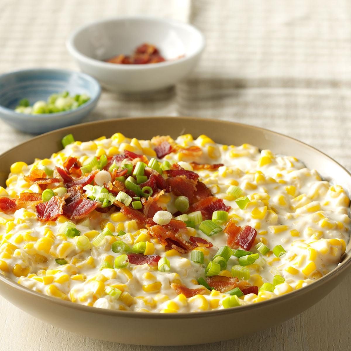 Creamed corn topped with bacon and green onions fills a bowl on a table, accompanied by two small dishes in the background.