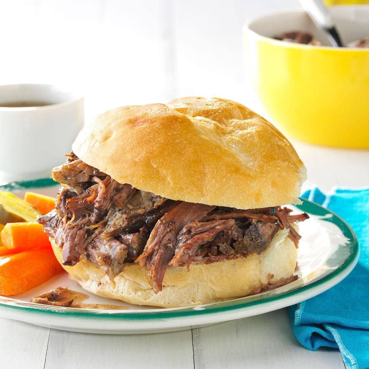 Sandwich filled with shredded beef sits on a plate alongside carrot sticks and pickles, with a bowl and sauce cup in the background.