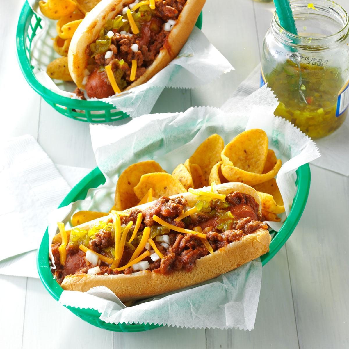 Chili cheese hot dogs topped with relish sit in green baskets, accompanied by corn chips, on a light wooden table with a relish jar nearby.