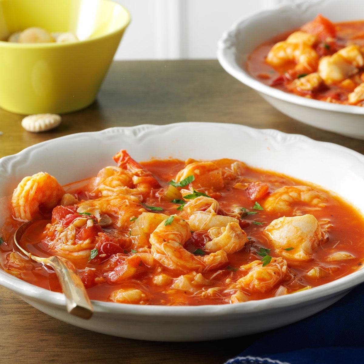 Seafood stew fills a white bowl, showcasing shrimp in a rich, red tomato broth; a yellow bowl and crackers are nearby on the table.