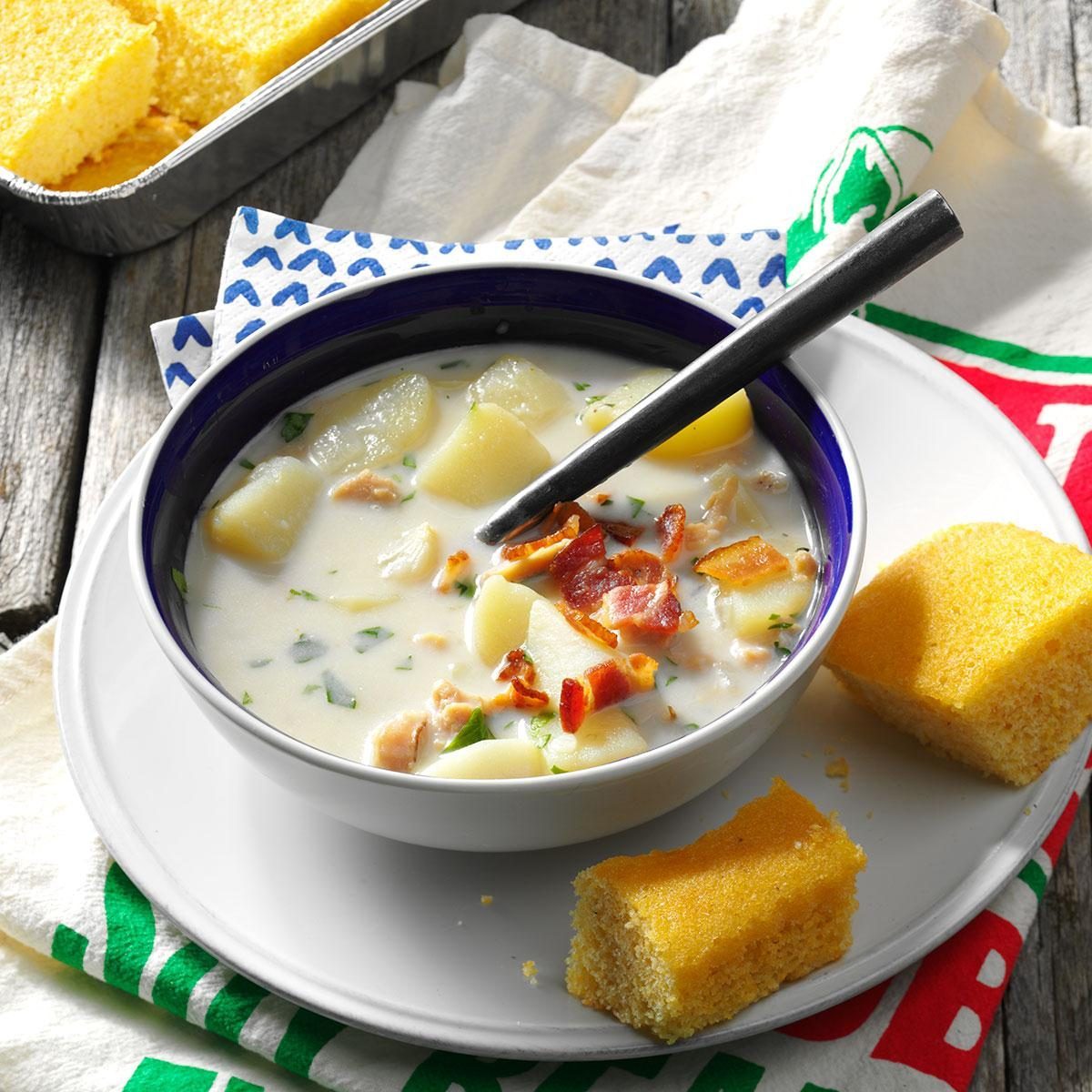 A soup bowl contains creamy potato soup with bacon, next to cornbread, on a rustic table setting.