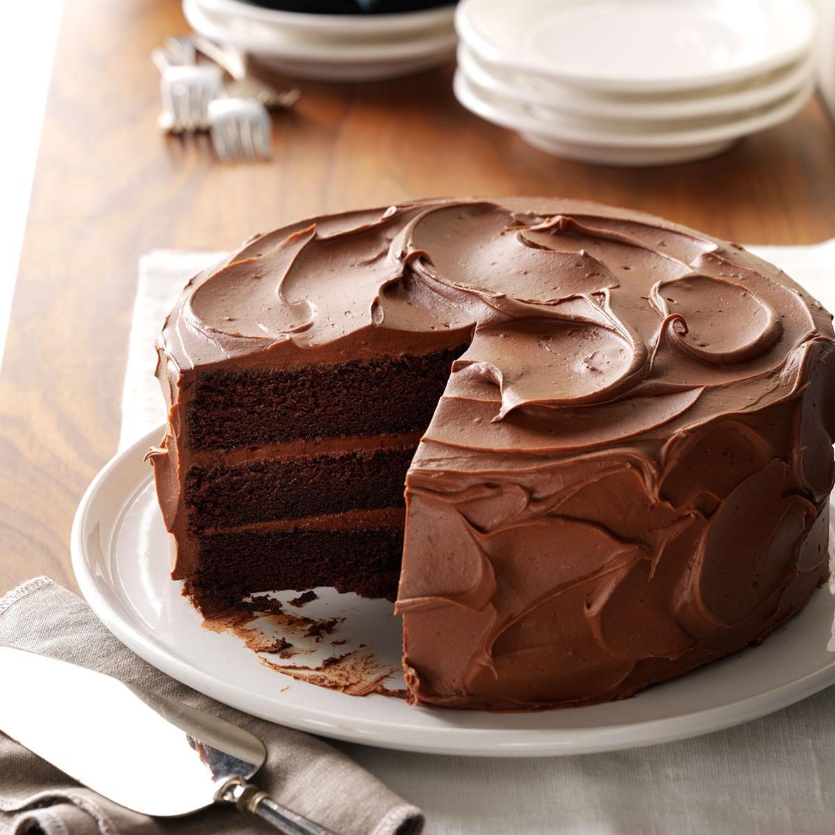 Chocolate cake, sliced, sits on a white plate with rich frosting. Plates and forks are stacked nearby on a wooden table.