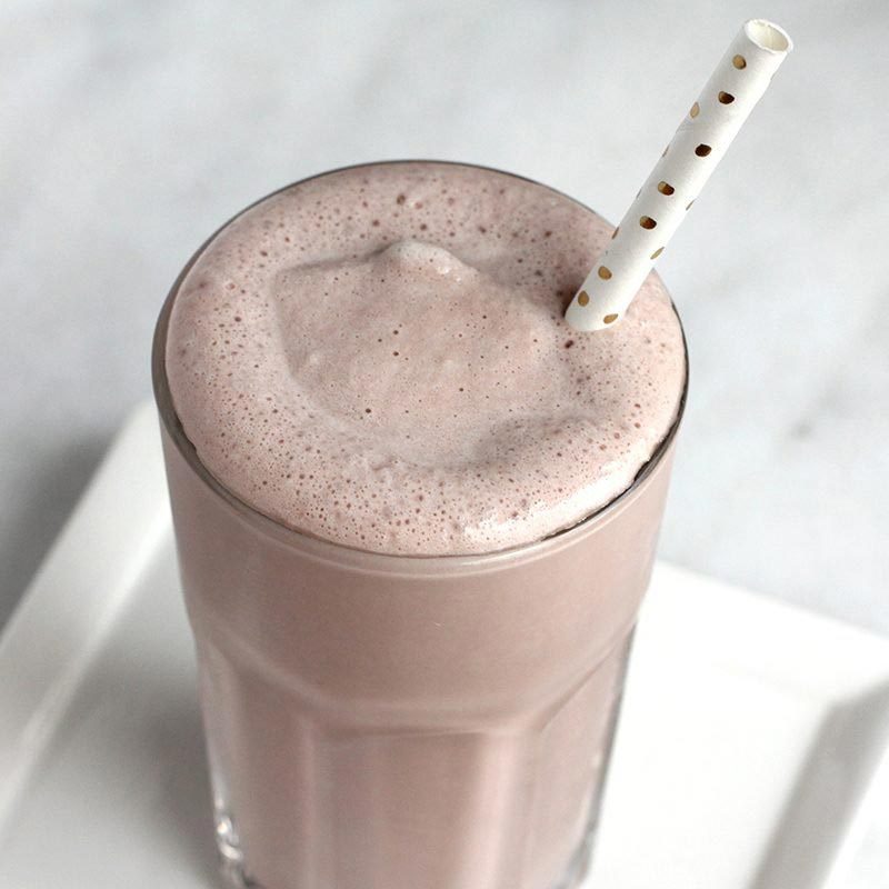 A frothy chocolate milkshake sits in a glass with a polka-dot straw, placed on a light surface.