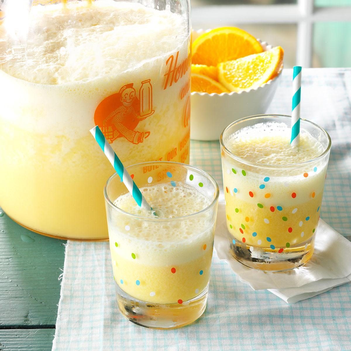 Two glasses filled with frothy orange drink, each with a blue striped straw, sit on a checkered cloth beside a bowl of orange slices.